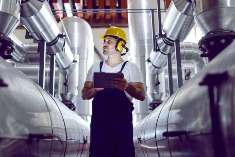 A man wearing a hard hat and protective ear muffs surveys equipment in an industrial plant.